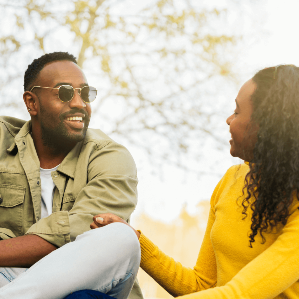 A smiling couple sitting outdoors, enjoying a sunny day. The man is wearing sunglasses and a green jacket, while the woman is dressed in a yellow sweater. The photo showcases the benefits of professional dating coaching in Orlando, highlighting natural connections and confidence in dating.