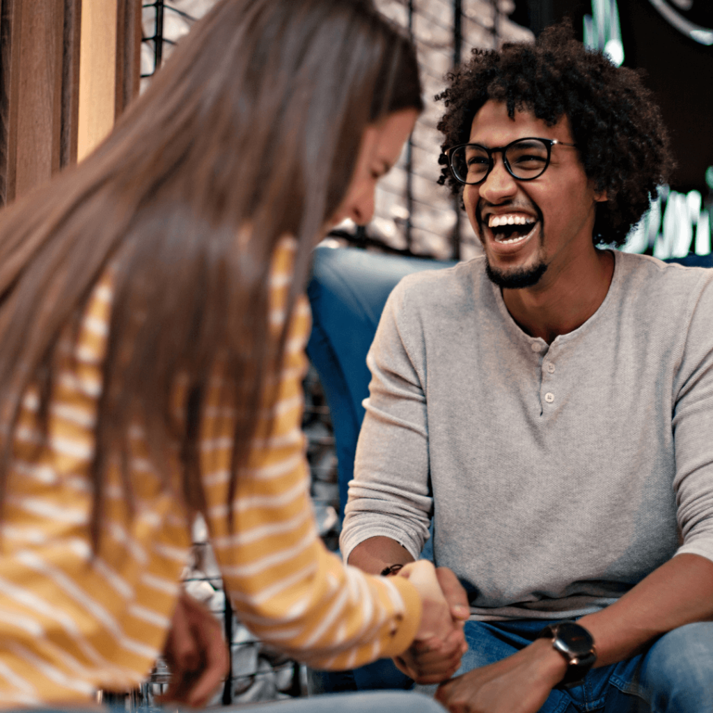 A joyful man in glasses and a gray sweater, shaking hands with a woman in a casual setting. They share a laugh, capturing the essence of building rapport and connection through dating coaching. The image promotes advanced dating techniques and confidence-boosting sessions in Orlando.