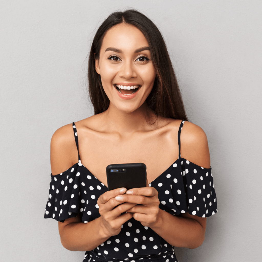 A woman in a polka dot dress smiling excitedly while holding a smartphone, representing the joy and success of receiving a positive message or match in online dating, thanks to Orlando Date Coach's guidance.
