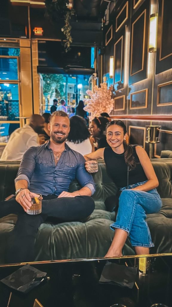 Ty Temmel, Orlando's top dating coach, enjoying a relaxed evening out at a stylish lounge with a woman, both holding drinks and smiling. The image reflects the social success and confidence gained through his dating coaching services.