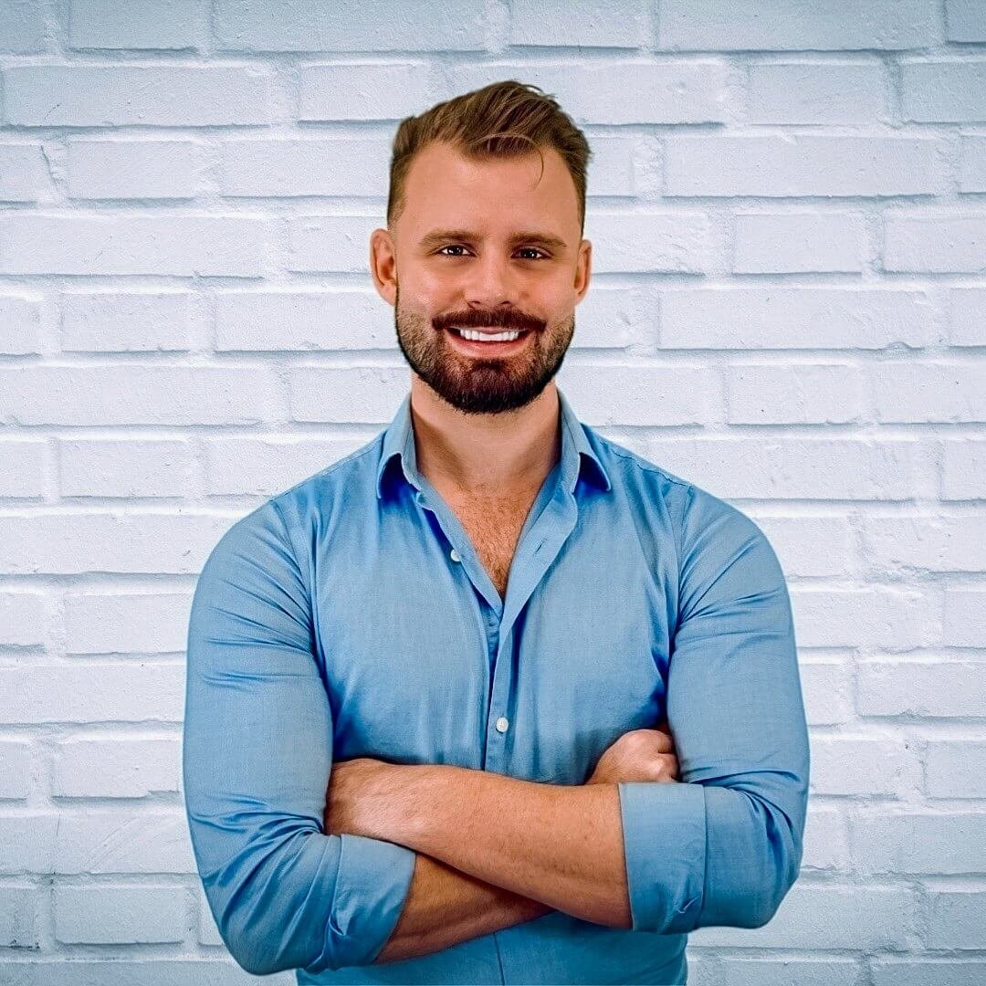 Ty Temmel, Orlando's premier dating coach, smiling confidently in a blue shirt with arms crossed in front of a white brick wall. The image represents the approachable and professional nature of his dating coaching services.