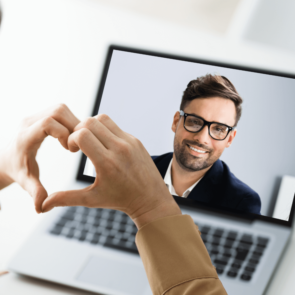 A person forming a heart shape with their hands while video chatting with a smiling man on a laptop screen. The image highlights the success of virtual dating and the guidance provided by Orlando Date Coach.