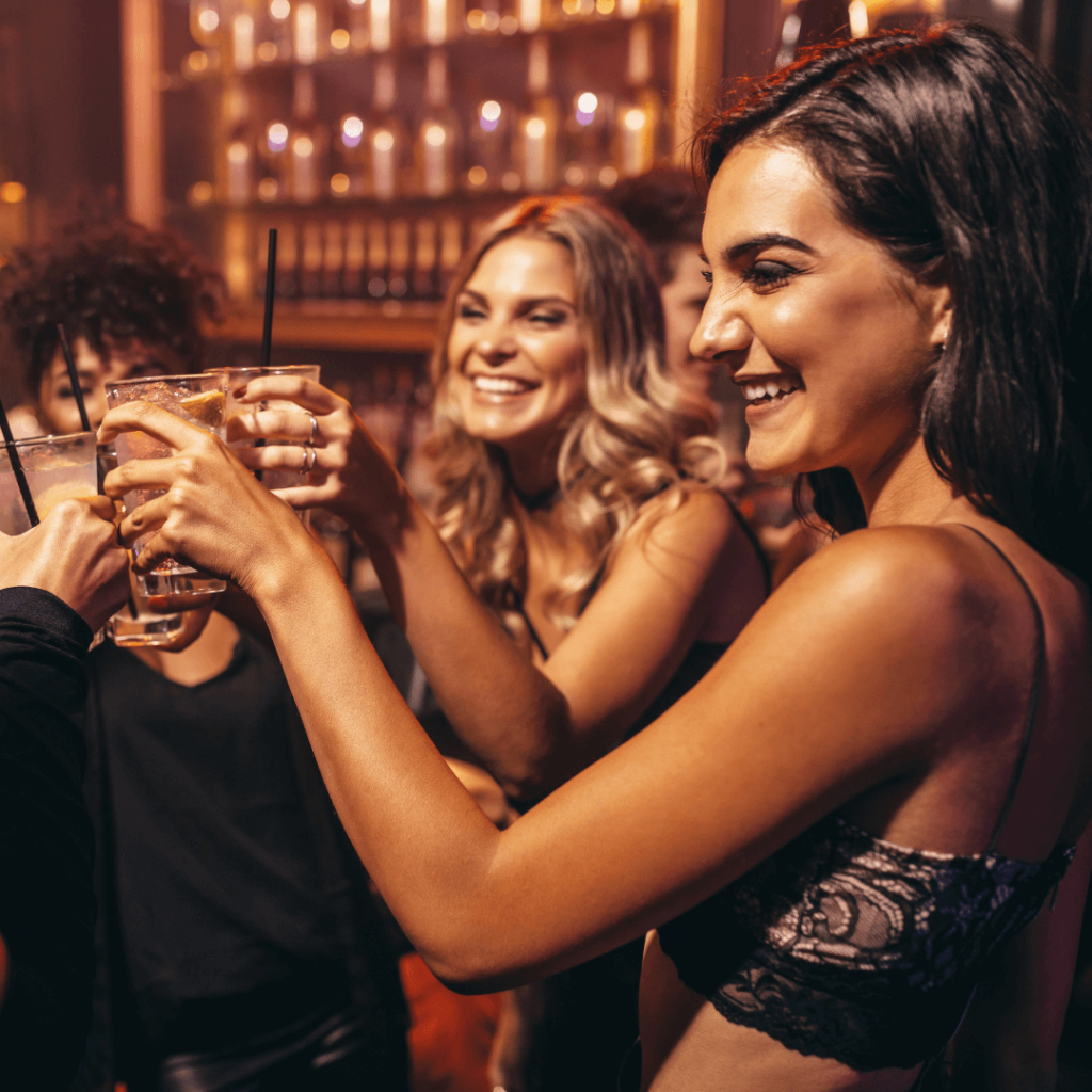 A group of smiling women in a lively bar setting, raising their glasses for a toast. The image represents the social and dating scene in Orlando, highlighting the benefits of group coaching and social skills improvement offered by Orlando Date Coach.